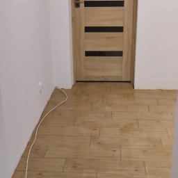 Yurii Drohomyretskyi KUB - Jasne, drewnopodobne płytki podłogowe ułożone w nowym wnętrzu z drewnianymi drzwiami z czarnymi, poziomymi paskami. Widoczny fragment białej ściany z kablem.