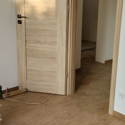 Yurii Drohomyretskyi KUB - Otwarta, drewniana, wewnętrzna ościeżnica z zamontowanymi drzwiami w odcieniach beżu, widok na podłogę z paneli imitujących drewno, gniazdko elektryczne.