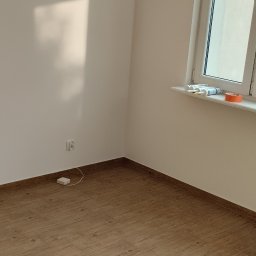 Yurii Drohomyretskyi KUB - Jasne, wykończone wnętrze z panelami podłogowymi imitującymi drewno. Widoczny fragment ściany z gniazdkiem i okno z akcesoriami na parapecie.