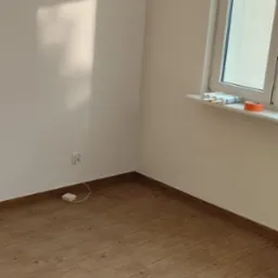 Jasne, wykończone wnętrze z panelami podłogowymi imitującymi drewno. Widoczny fragment ściany z gniazdkiem i okno z akcesoriami na parapecie.