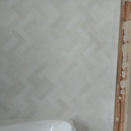 Yurii Drohomyretskyi KUB - Ściana w łazience wyłożona jasnymi płytkami w jodełkę, z widoczną białą wanną i fragmentem ościeżnicy. Minimalistyczny design wnętrza, w trakcie prac wykończeniowych.