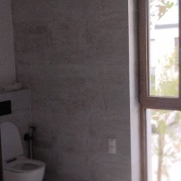 Yurii Drohomyretskyi KUB - Jasna łazienka z toaletą i oknem w drewnianej ramie. Ściany i podłoga wyłożone płytkami w odcieniach szarości. Widoczne wystające kable z sufitu.