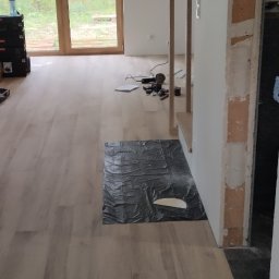 Yurii Drohomyretskyi KUB - Jasne panele podłogowe ułożone w nowym domu, zabezpieczone folią. W tle widoczne narzędzia i okno z widokiem na zieleń.