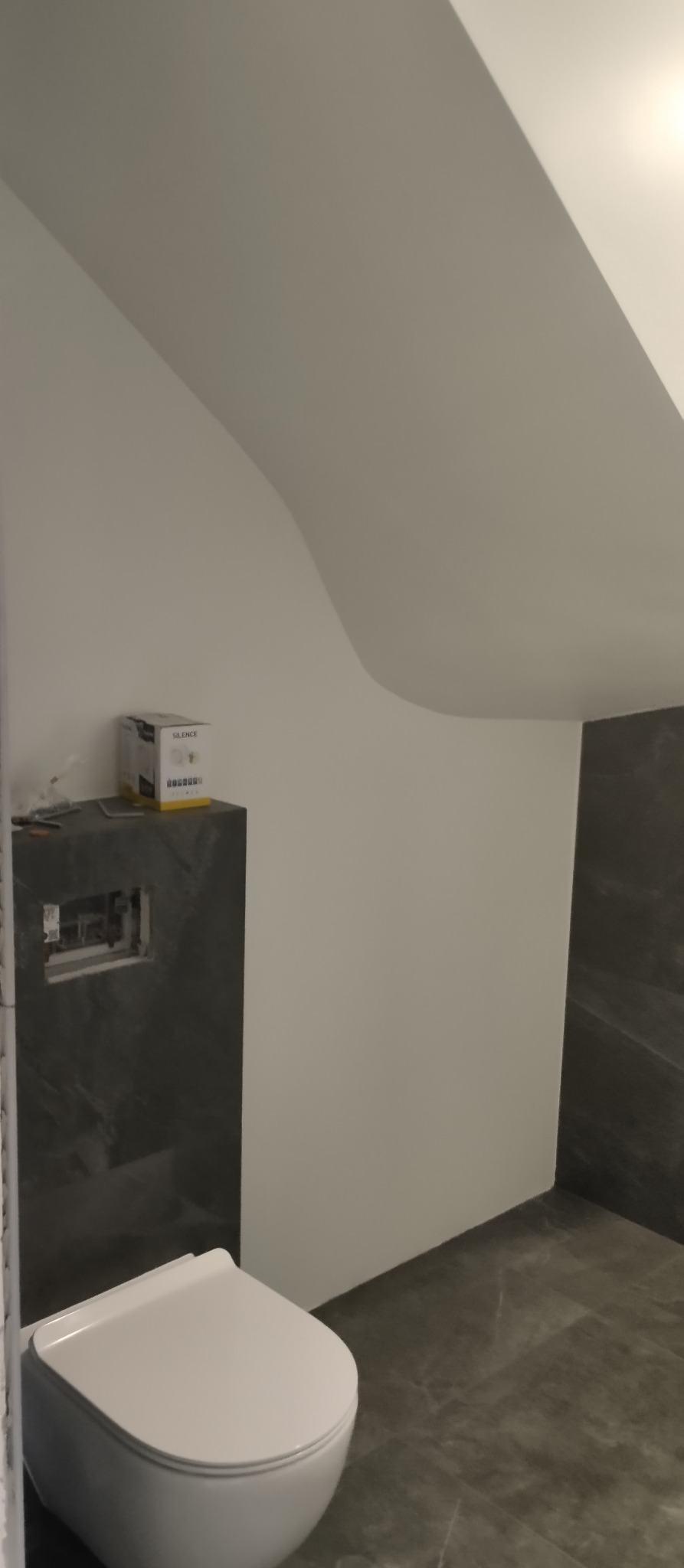 Nowoczesna toaleta podwieszana w łazience z szarą marmurową okładziną i białą ścianą pod skosem. Widoczna instalacja podtynkowa i pudełko na blacie.