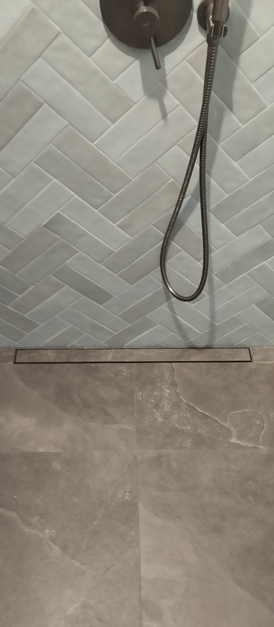 Nowoczesna łazienka: jasne płytki ułożone w jodełkę na ścianie, szara podłoga i minimalistyczna armatura prysznicowa z odpływem liniowym.