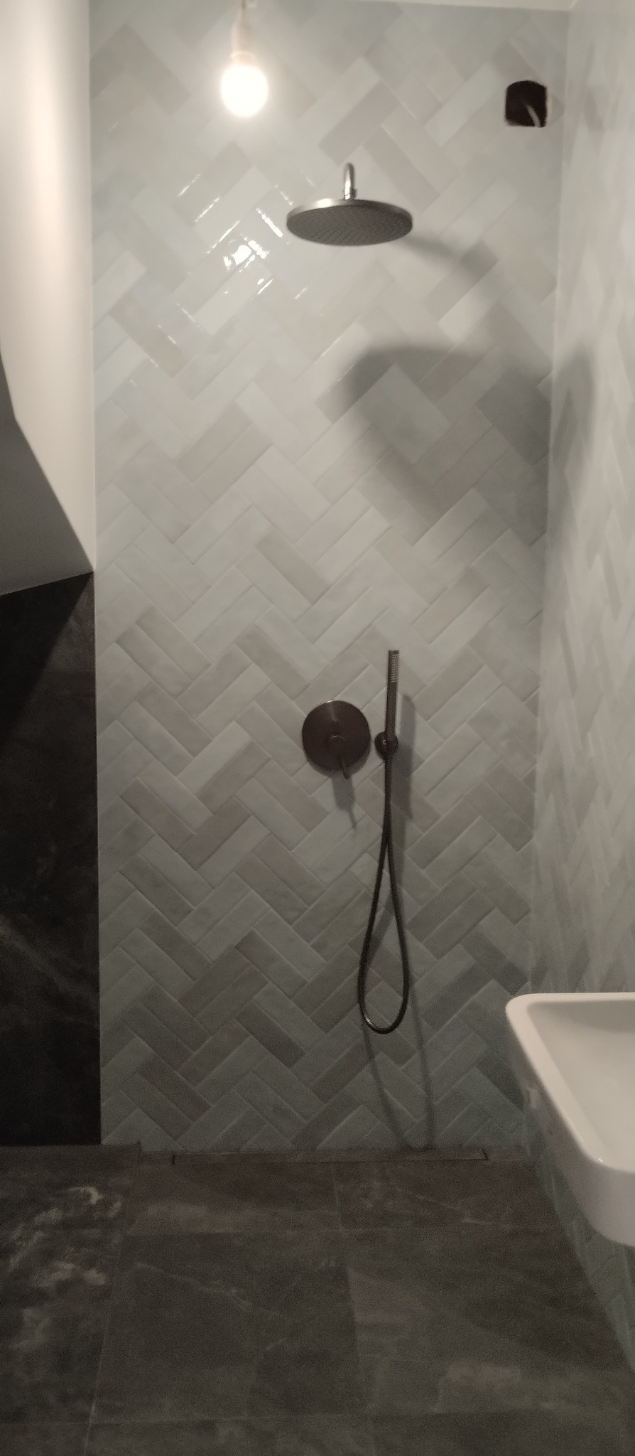 Nowoczesna łazienka po remoncie: Ściany wyłożone jasnymi, błyszczącymi płytkami w jodełkę, deszczownica i bateria w kolorze brązu, ciemna podłoga z płytek imitujących kamień.