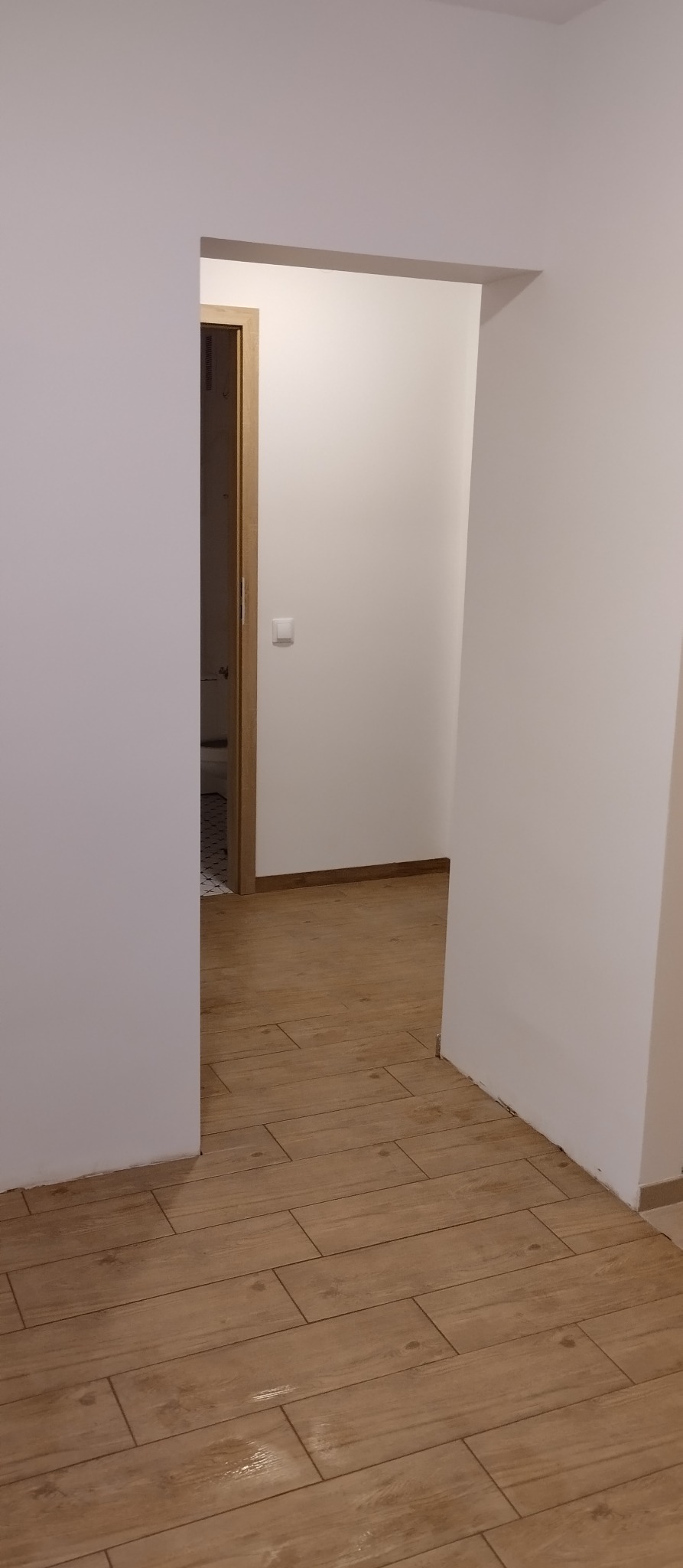Beżowe płytki podłogowe imitujące drewno w korytarzu z białymi ścianami. Widok na otwarte drzwi z drewnianą ościeżnicą i fragmentem podłogi w kolejnym pomieszczeniu.