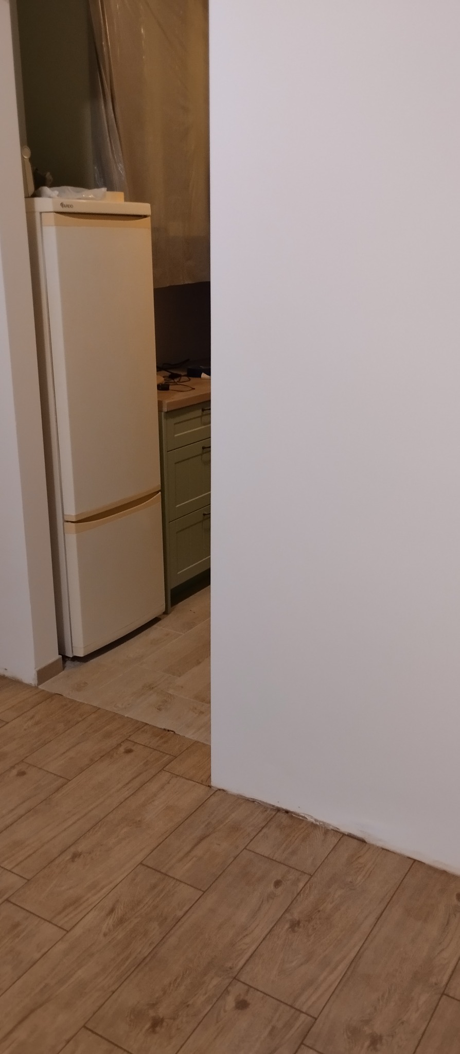 Ukończona kuchnia z kremową lodówką i drewnopodobną podłogą. Widok na zielone szafki kuchenne w tle. Świeżo pomalowana biała ściana po prawej stronie kadru.