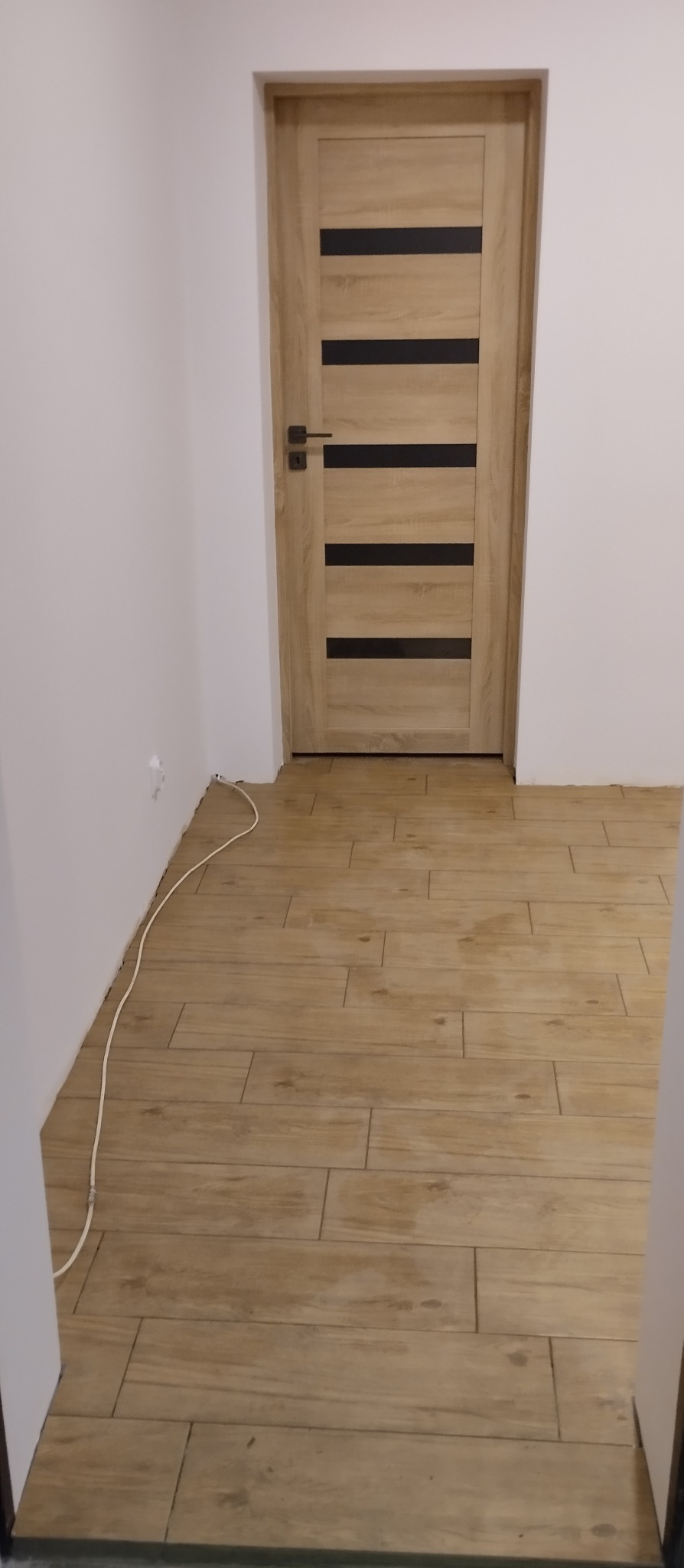 Jasne, drewnopodobne płytki podłogowe ułożone w nowym wnętrzu z drewnianymi drzwiami z czarnymi, poziomymi paskami. Widoczny fragment białej ściany z kablem.