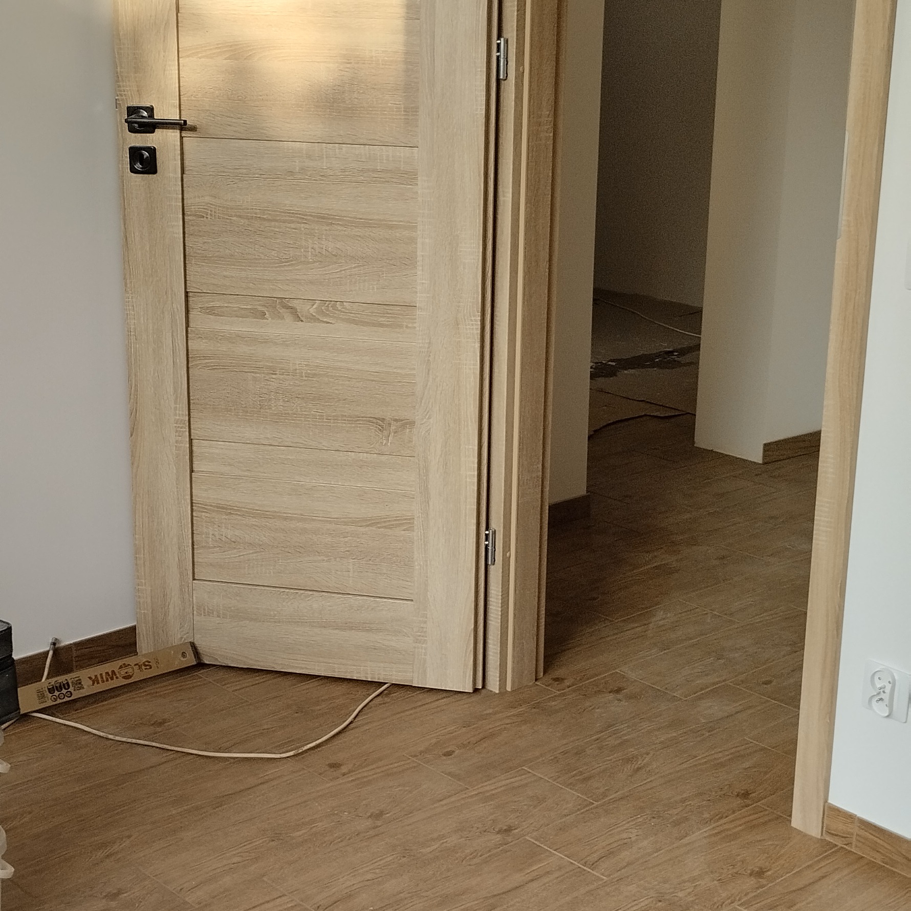 Otwarta, drewniana, wewnętrzna ościeżnica z zamontowanymi drzwiami w odcieniach beżu, widok na podłogę z paneli imitujących drewno, gniazdko elektryczne.