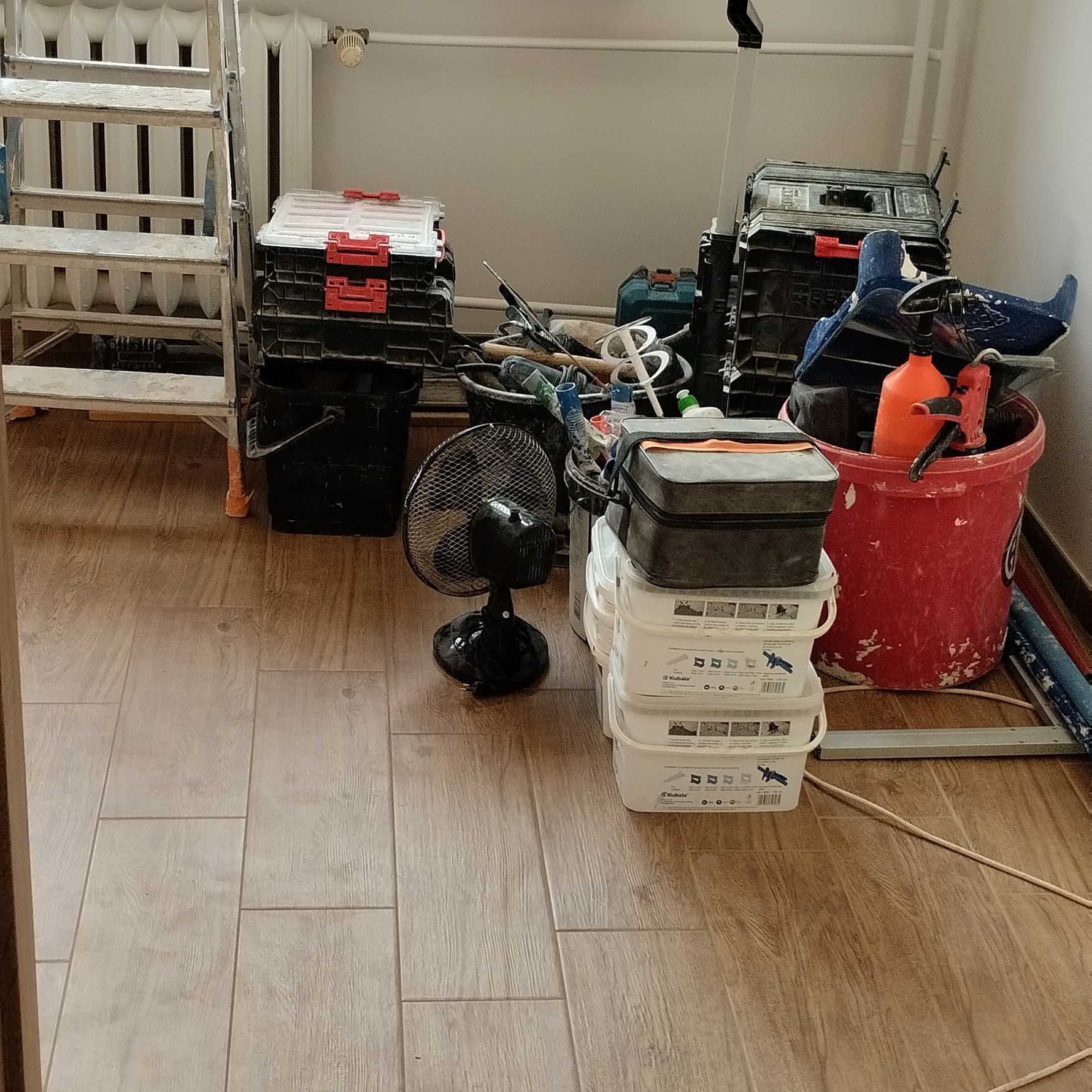 Ułożone płytki podłogowe w neutralnym odcieniu, otoczone narzędziami i akcesoriami budowlanymi: drabina, skrzynki narzędziowe, wiadra z farbą, wentylator.