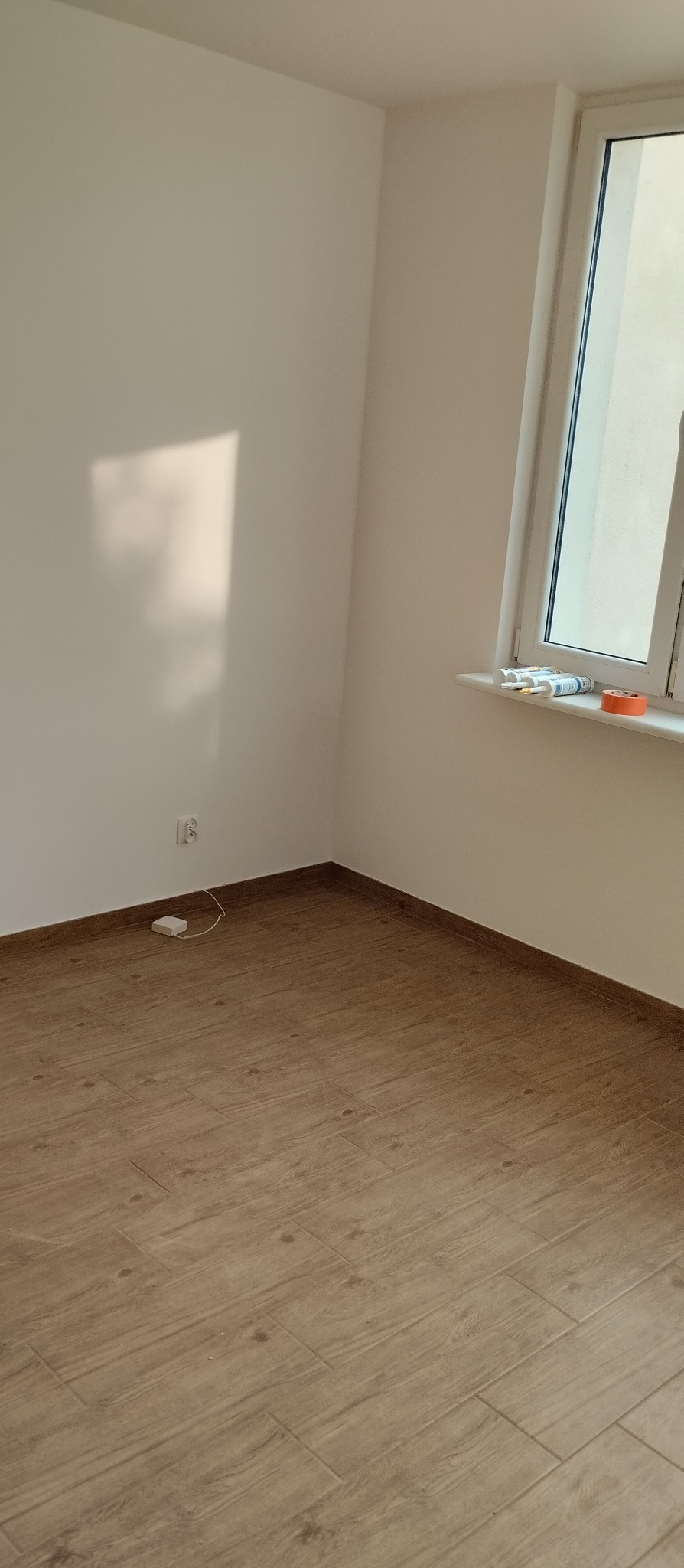 Jasne, wykończone wnętrze z panelami podłogowymi imitującymi drewno. Widoczny fragment ściany z gniazdkiem i okno z akcesoriami na parapecie.