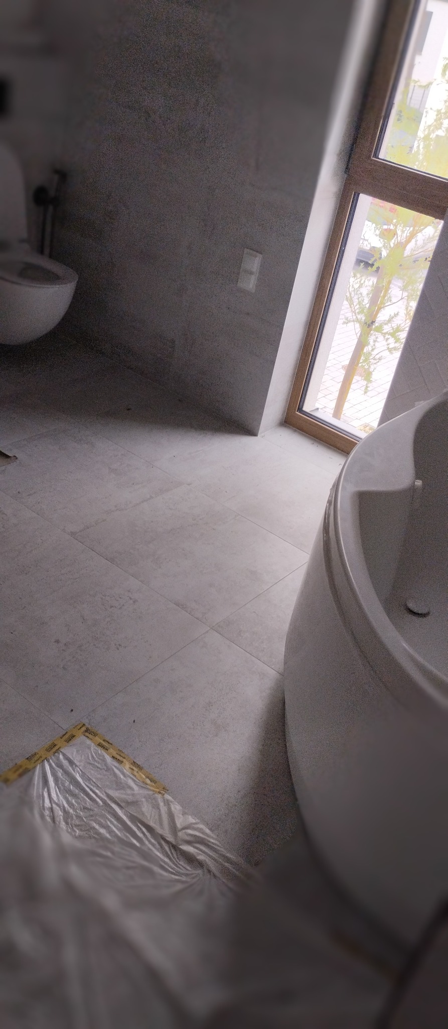 Szara łazienka w trakcie prac: widoczna wanna, toaleta i zabezpieczona folią podłoga z szarych płytek. Okno z drewnianą ramą wpuszcza naturalne światło.