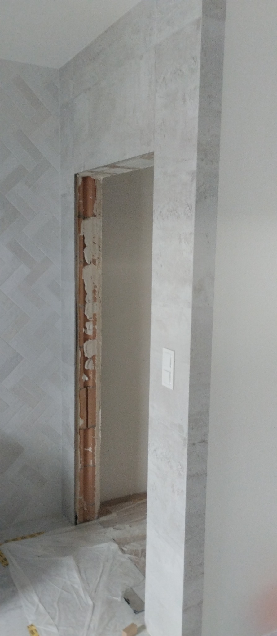 Wykończenie otworu drzwiowego płytkami imitującymi kamień, widoczna ściana z wzorem jodełki i zabezpieczona podłoga w trakcie prac.