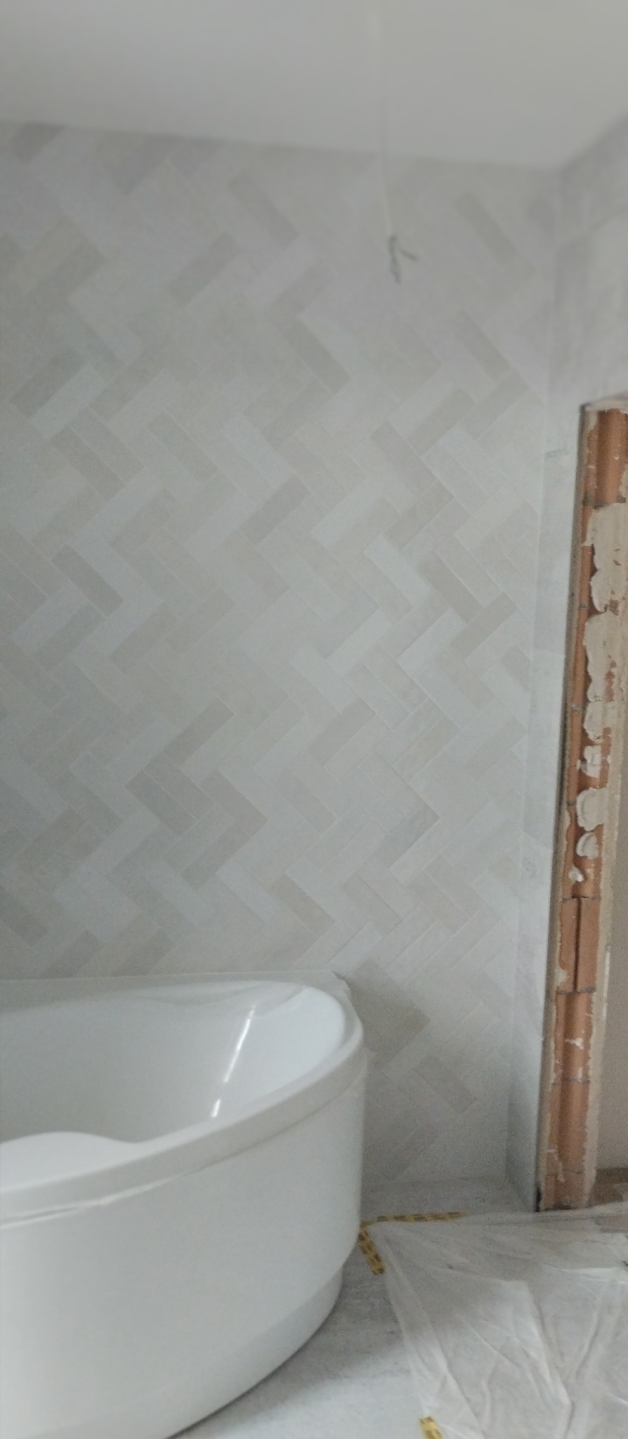 Ściana w łazience wyłożona jasnymi płytkami w jodełkę, z widoczną białą wanną i fragmentem ościeżnicy. Minimalistyczny design wnętrza, w trakcie prac wykończeniowych.