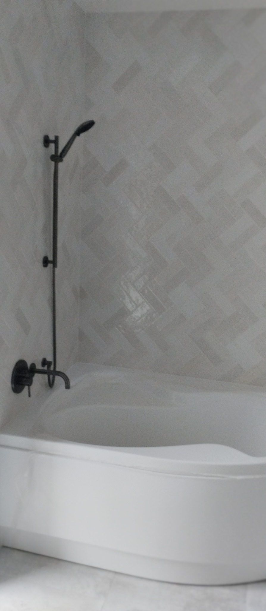 Biała wanna z czarną armaturą i prysznicem na tle ściany wyłożonej jasnymi płytkami w jodełkę, tworząc minimalistyczny i nowoczesny wystrój łazienki.