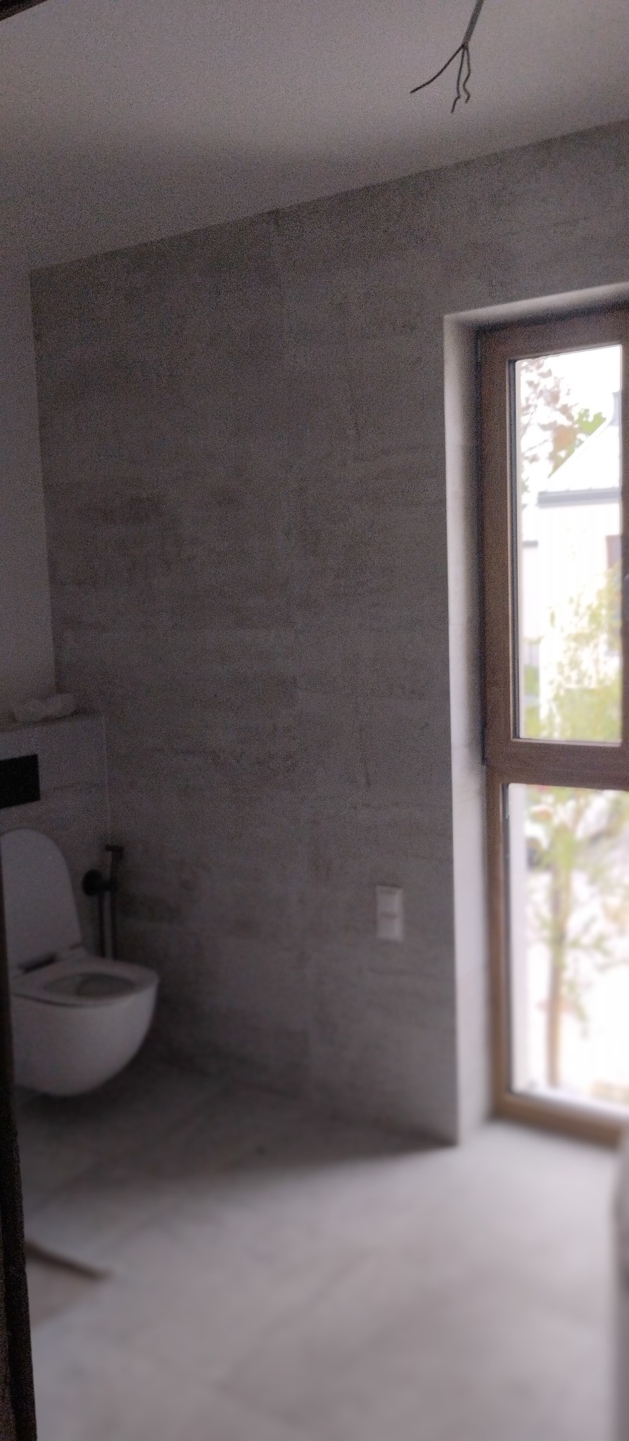 Jasna łazienka z toaletą i oknem w drewnianej ramie. Ściany i podłoga wyłożone płytkami w odcieniach szarości. Widoczne wystające kable z sufitu.