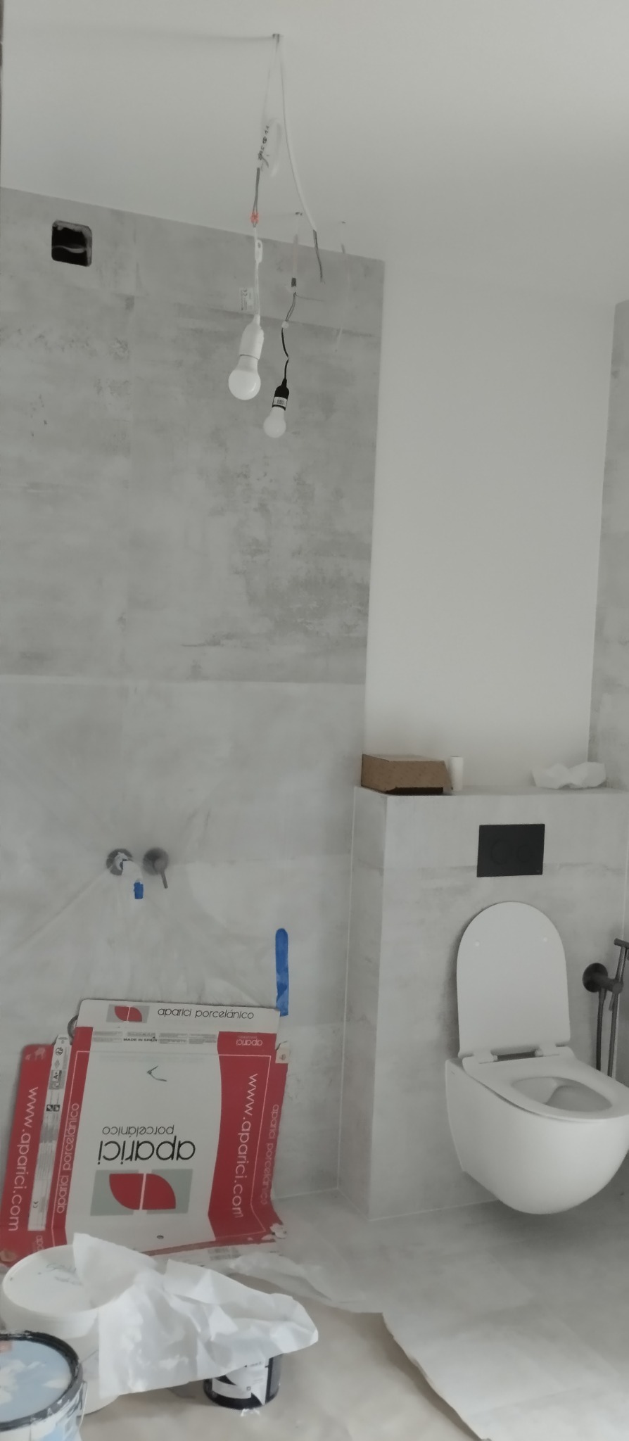 Remont łazienki w Warszawie: ułożone płytki imitujące beton, podwieszana toaleta. Widoczne kartony po płytkach i prowizoryczne oświetlenie.