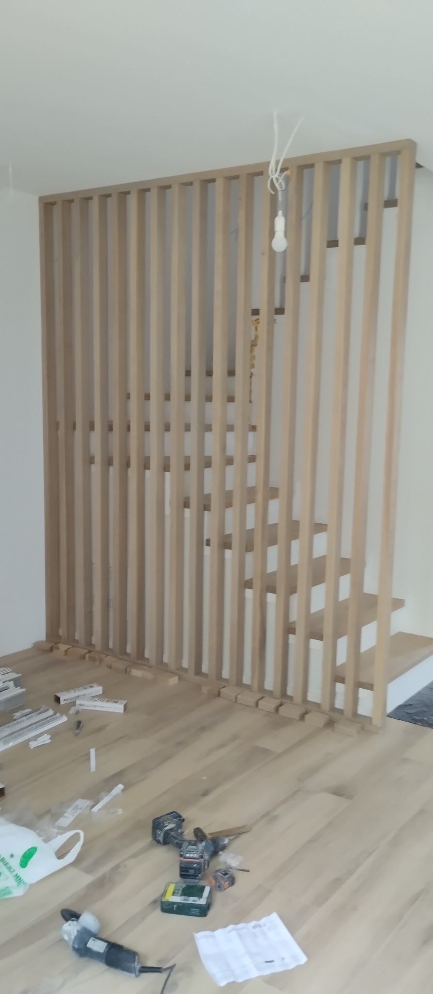 Montaż drewnianej ażurowej ścianki działowej przy schodach. Widoczne narzędzia i resztki materiałów na podłodze z paneli. Surowy stan wykończenia wnętrza.