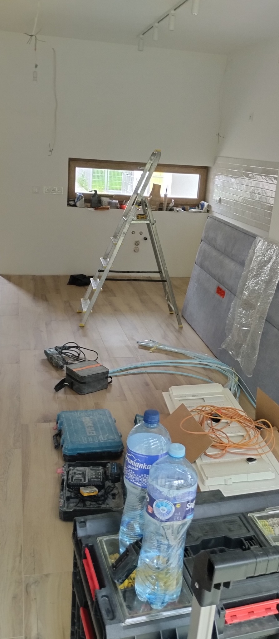 Wnętrze w trakcie remontu: drabina, narzędzia, kable, listwy przypodłogowe i materiały budowlane na podłodze. Widoczne punkty oświetleniowe na suficie.