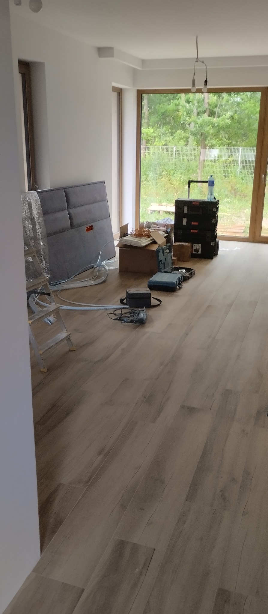 Jasne panele podłogowe w nowym wnętrzu, widoczne narzędzia i materiały przygotowane do montażu. Duże okno z widokiem na zieleń w tle.