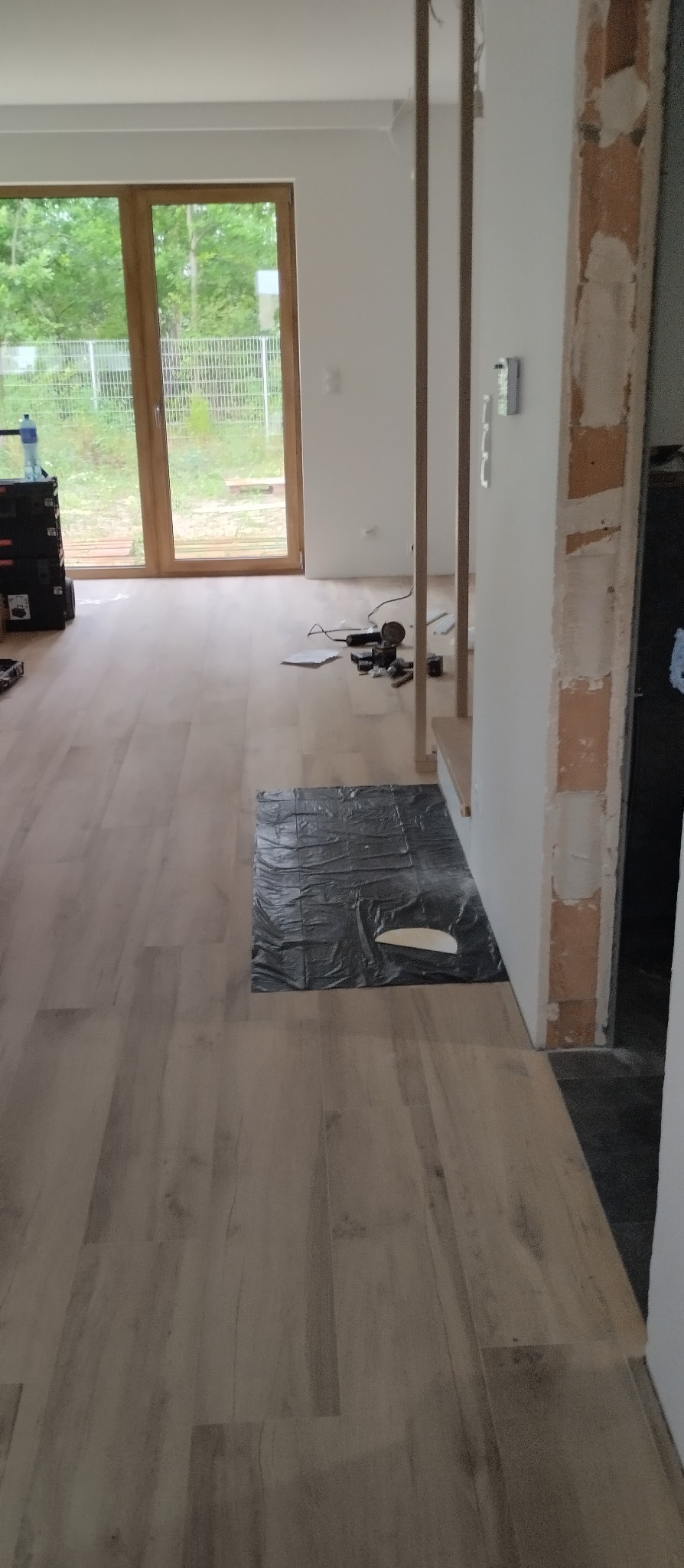 Jasne panele podłogowe ułożone w nowym domu, zabezpieczone folią. W tle widoczne narzędzia i okno z widokiem na zieleń.