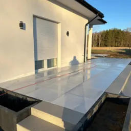 Białe płytki tarasowe ułożone na zewnątrz budynku z szarymi schodami i donicami, widoczne drzewa w tle.