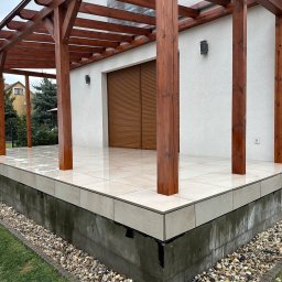 TarasCraft- Tarasy wentylowane - Nowoczesny taras z jasnych, błyszczących płytek, z drewnianą pergolą i widokiem na ogród, wykończony kamieniami ozdobnymi przy podstawie.