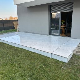 TarasCraft- Tarasy wentylowane - Nowoczesny taras wyłożony jasnymi, kwadratowymi płytkami, przylegający do szarego budynku z otwartymi drzwiami balkonowymi i widocznymi tulipanami w wazonie wewnątrz.