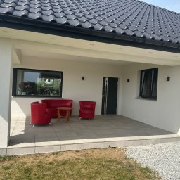 TarasCraft- Tarasy wentylowane - Zadaszony taras wyłożony jasnymi, kwadratowymi płytkami, z kompletem czerwonych mebli wypoczynkowych i małym drewnianym stolikiem; widok na dom z ciemną dachówką i fragment ogrodu z białym żwirem.