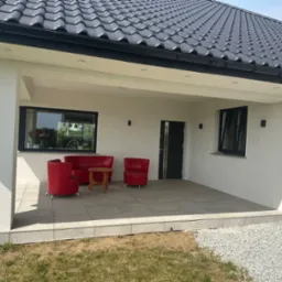 Zadaszony taras wyłożony jasnymi, kwadratowymi płytkami, z kompletem czerwonych mebli wypoczynkowych i małym drewnianym stolikiem; widok na dom z ciemną dachówką i fragment ogrodu z białym żwirem.