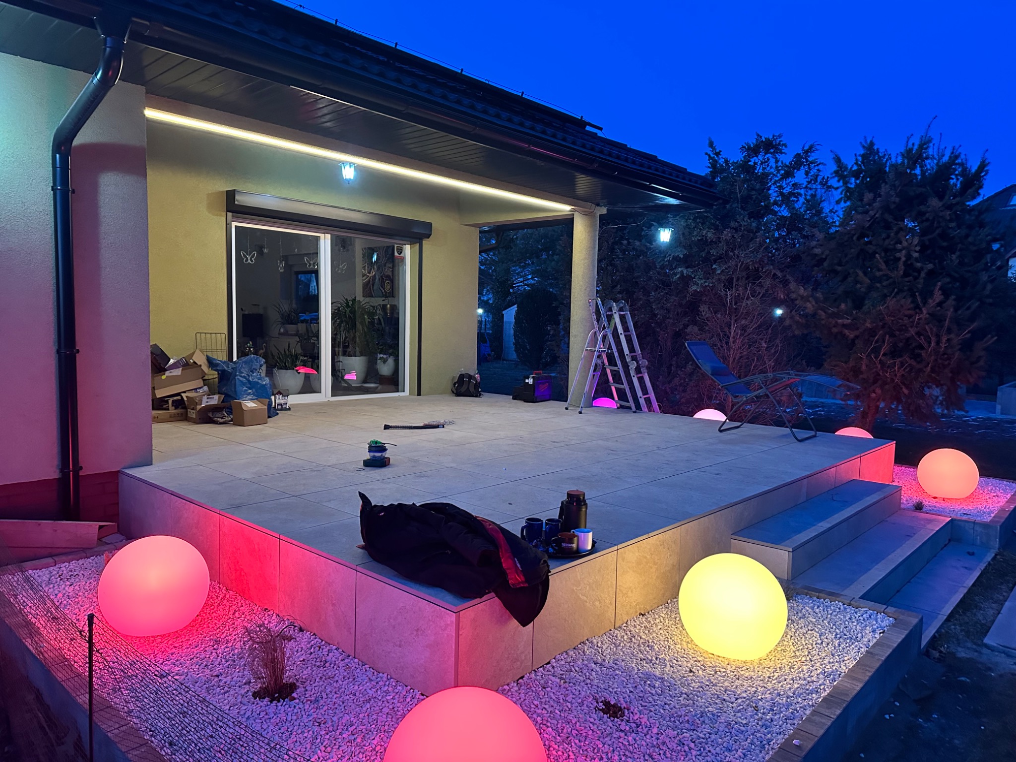 Podświetlony taras z kamienną nawierzchnią, widoczne kule świetlne na żwirowym podłożu, drabina i akcesoria budowlane sugerujące prace wykończeniowe.