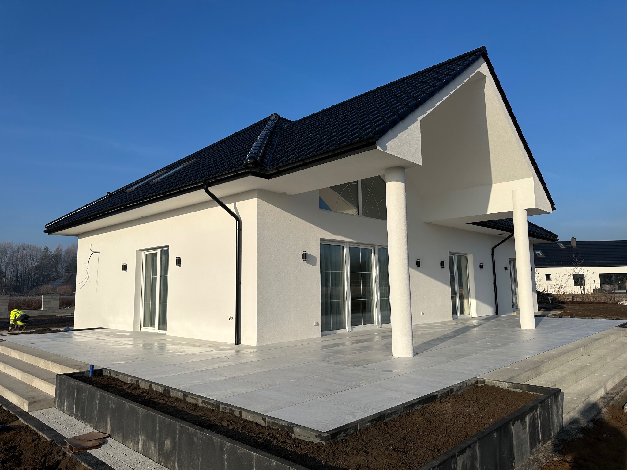 Nowoczesny, jasny taras z dużych, szarych płyt, przylegający do białego domu z czarnym dachem, widoczne prace wykończeniowe w ogrodzie.
