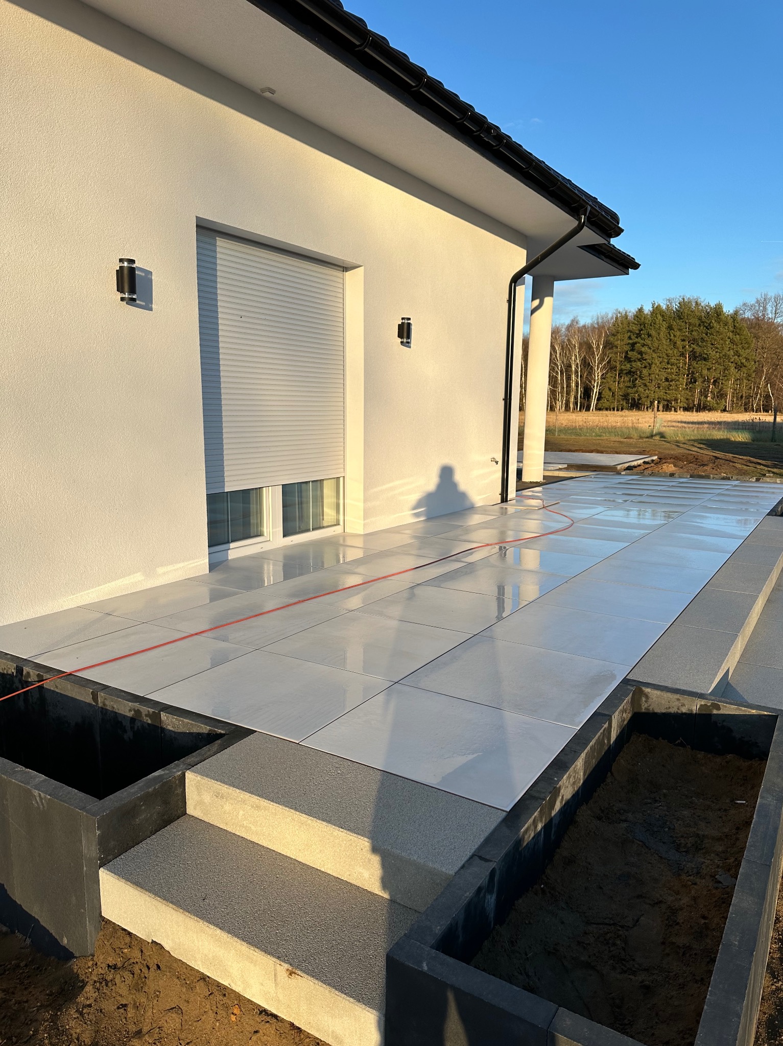 Białe płytki tarasowe ułożone na zewnątrz budynku z szarymi schodami i donicami, widoczne drzewa w tle.