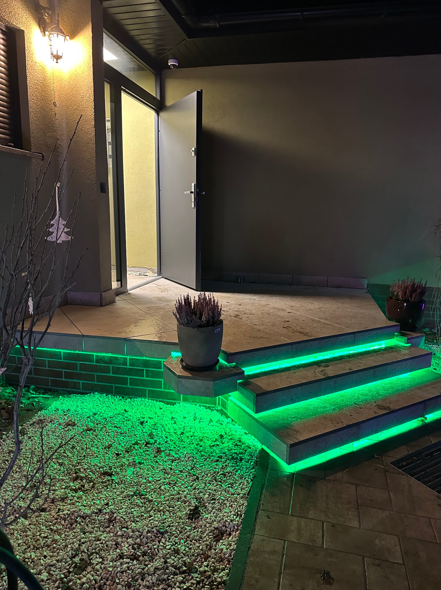 Oświetlony zielonymi LED-ami taras z kamiennymi schodami prowadzący do otwartych drzwi wejściowych, donica z wrzosem na pierwszym planie, żwirowe podłoże.