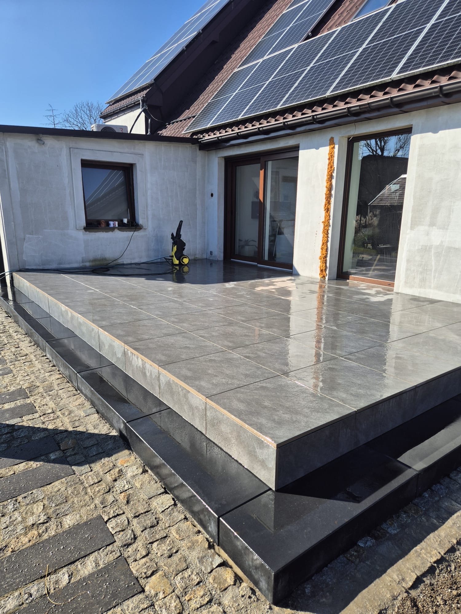 Nowoczesny taras z szarych płytek, z dwoma stopniami obłożonymi czarnym kamieniem, widoczny sprzęt do mycia ciśnieniowego, panele słoneczne na dachu budynku w tle.