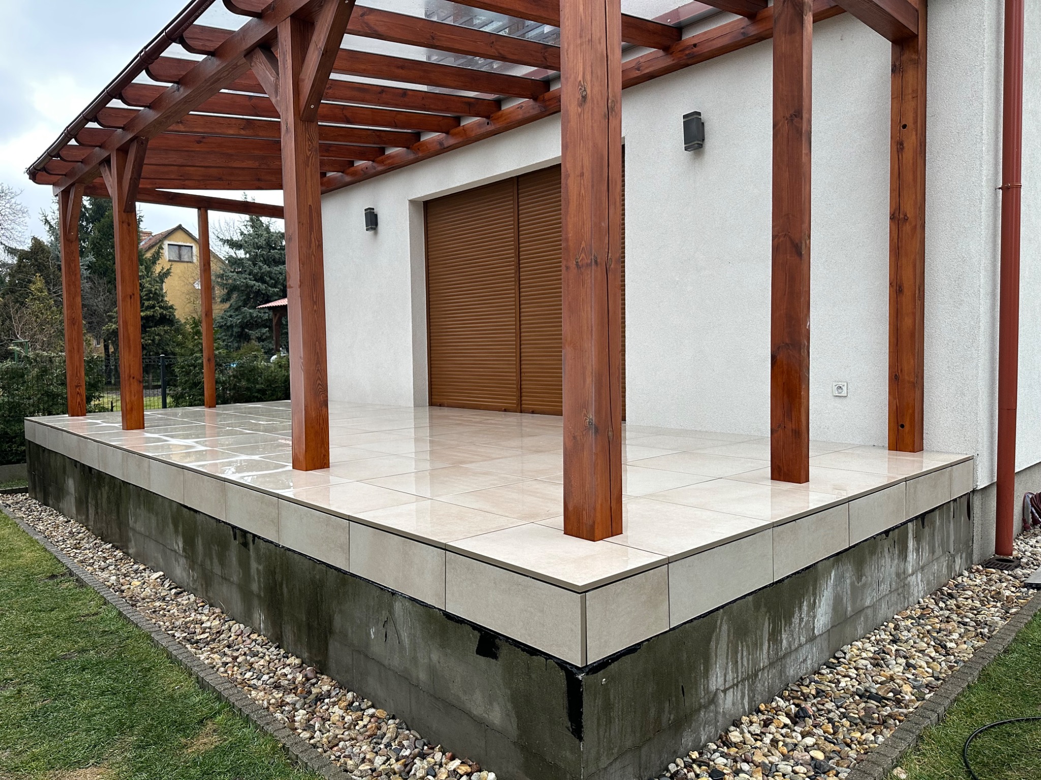 Nowoczesny taras z jasnych, błyszczących płytek, z drewnianą pergolą i widokiem na ogród, wykończony kamieniami ozdobnymi przy podstawie.