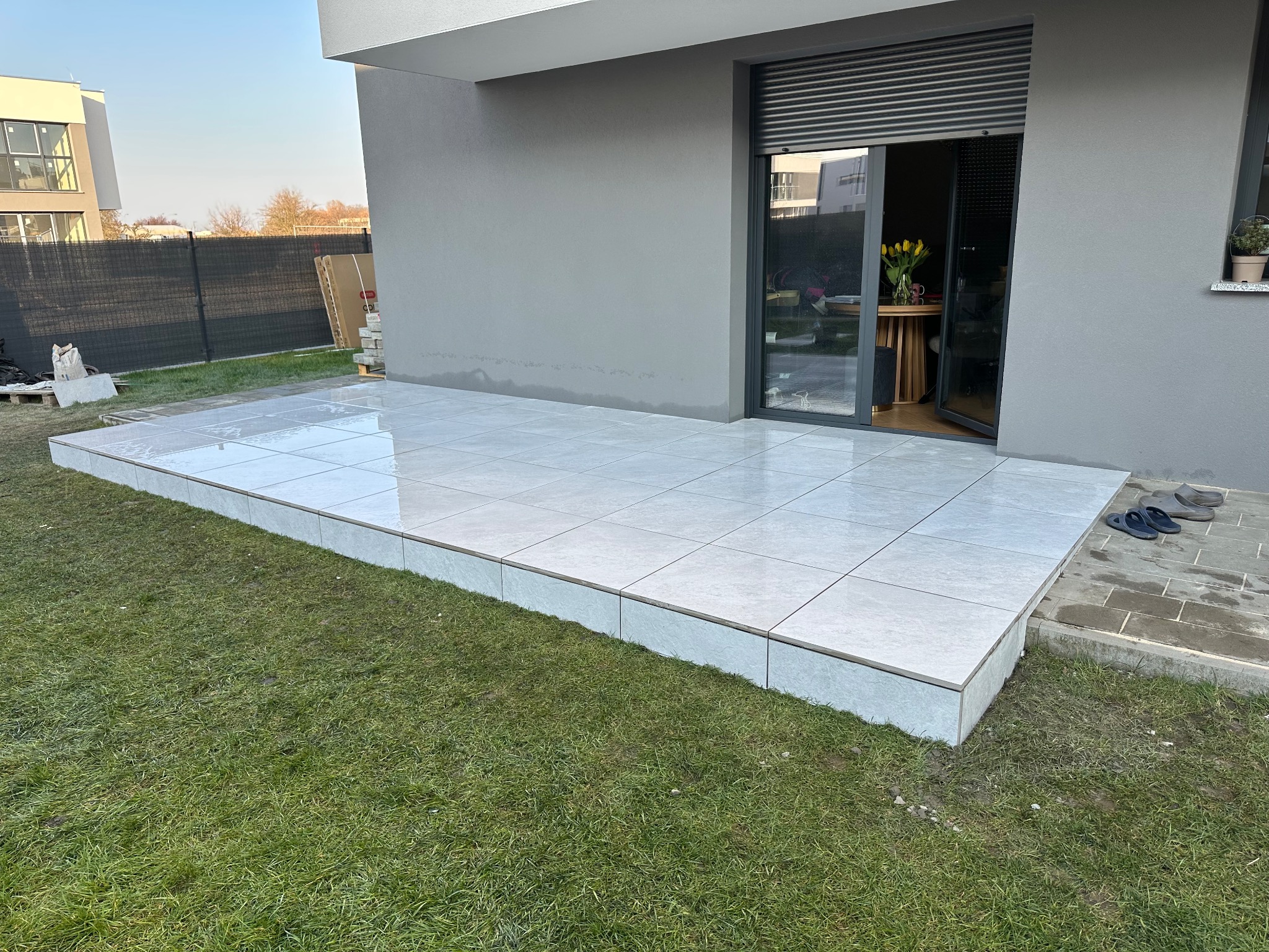 Nowoczesny taras wyłożony jasnymi, kwadratowymi płytkami, przylegający do szarego budynku z otwartymi drzwiami balkonowymi i widocznymi tulipanami w wazonie wewnątrz.