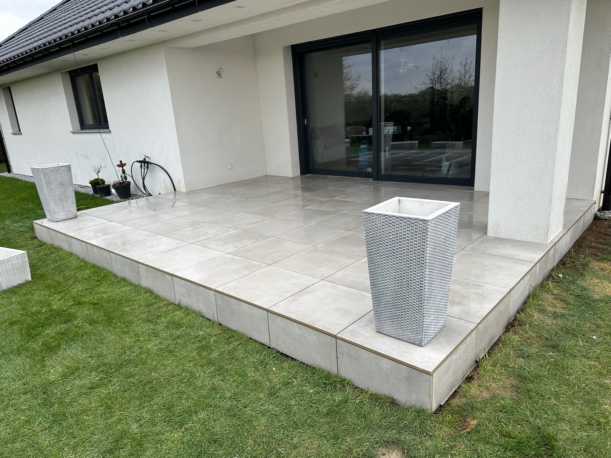 Nowoczesny taras kamienny z płyt w jasnych odcieniach szarości, z podwyższoną platformą i dekoracyjnymi donicami z tworzywa sztucznego, przylegający do białego budynku z dużymi przesuwnymi drzwiami...