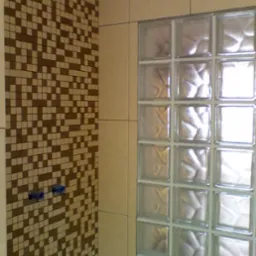 Ściana łazienki wyłożona mozaiką w odcieniach brązu i beżu, sąsiadująca z ścianą z luksferów, z widocznymi niebieskimi zaślepkami rur.