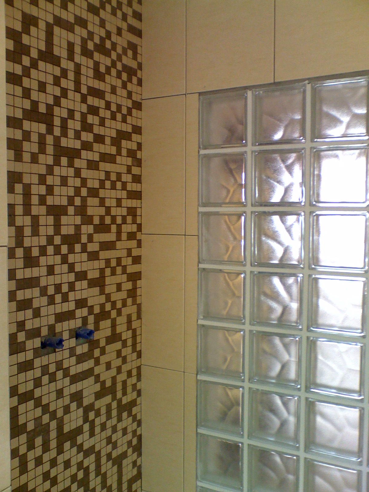 Ściana łazienki wyłożona mozaiką w odcieniach brązu i beżu, sąsiadująca z ścianą z luksferów, z widocznymi niebieskimi zaślepkami rur.