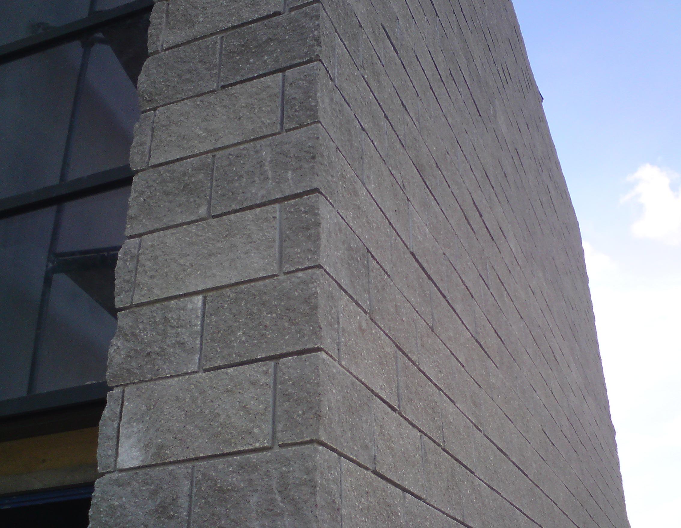 Szary narożnik budynku z bloków, widoczne okno z czarną ramą i fragment błękitnego nieba w tle.