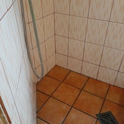 Tina - Kąt kabiny prysznicowej z brązowymi płytkami podłogowymi, odpływem i wężem prysznicowym zwisającym w rogu.