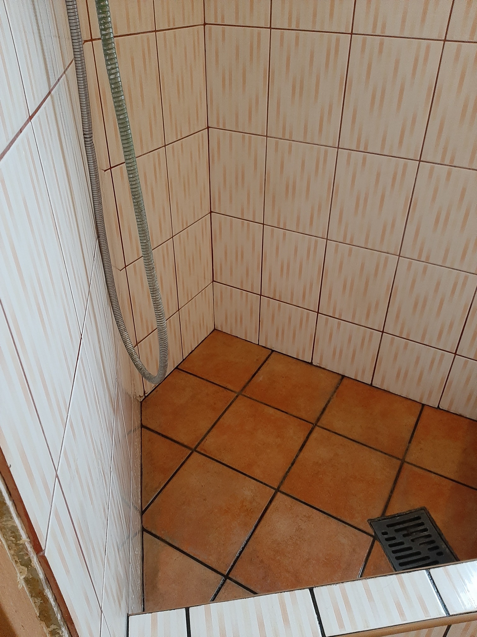 Kąt kabiny prysznicowej z brązowymi płytkami podłogowymi, odpływem i wężem prysznicowym zwisającym w rogu.