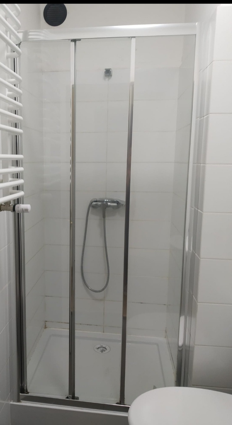 Kabina prysznicowa z przesuwnymi drzwiami, widoczne ślady użytkowania i osady na fugach, białe płytki i chromowana armatura w łazience.