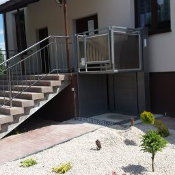 ERALIFT - Zewnętrzna platforma schodowa dla osób niepełnosprawnych, zamontowana przy budynku z jasną elewacją, obok schodów z metalową barierką, otoczona ozdobnym, białym żwirem i niską roślinnością.