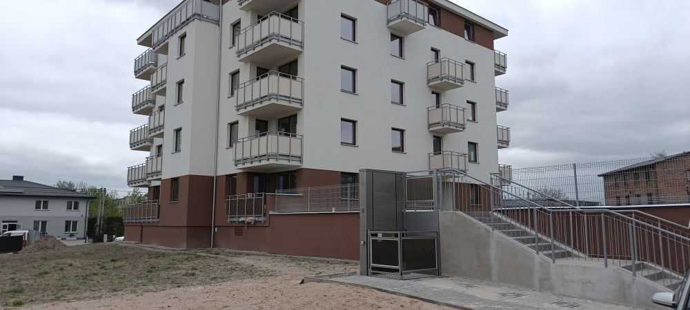 Zewnętrzny podnośnik platformowy dla osób niepełnosprawnych, zamontowany przy schodach wejściowych do nowoczesnego budynku wielorodzinnego z balkonami.