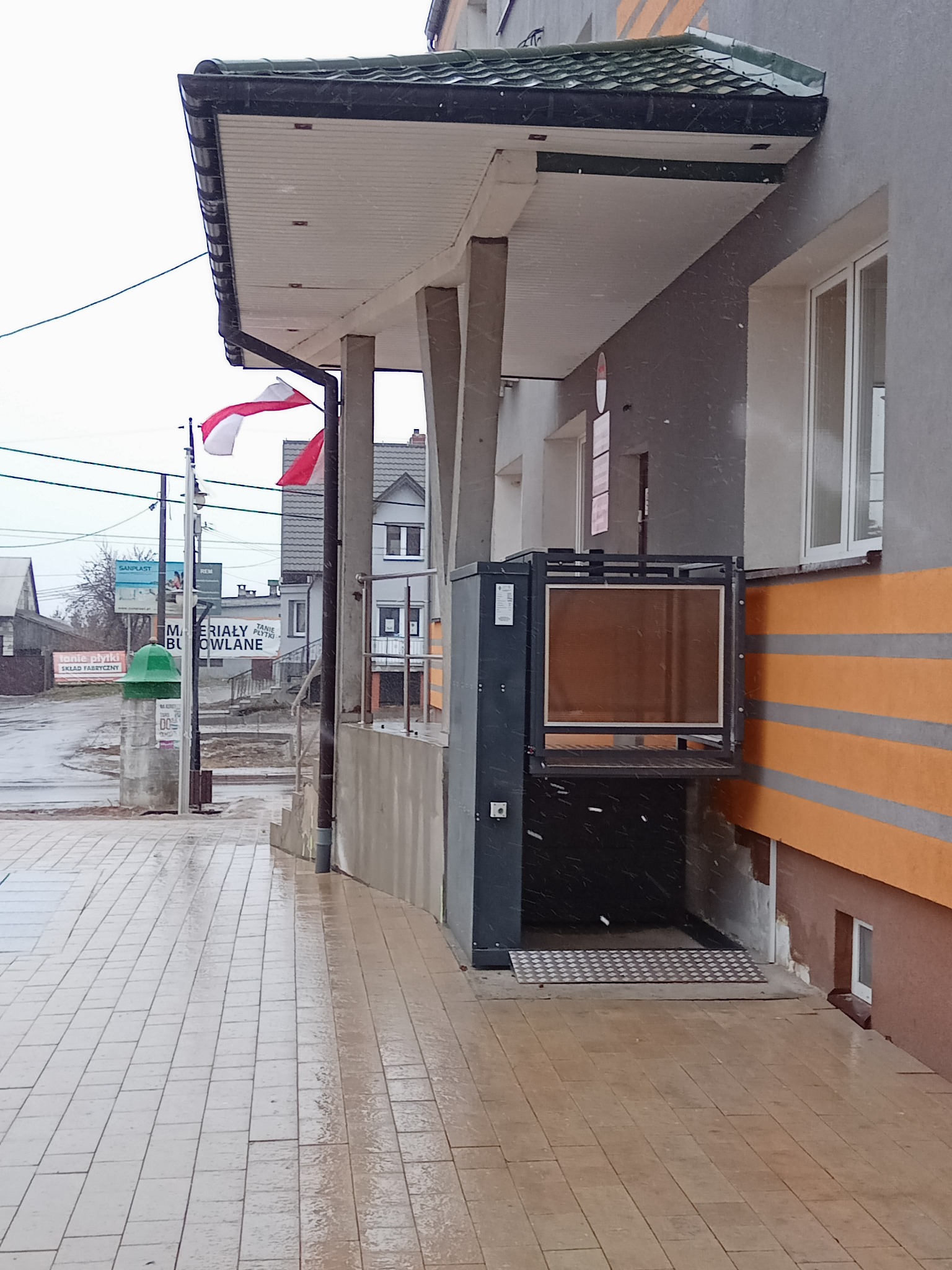 Zewnętrzna platforma schodowa dla osób niepełnosprawnych, zamontowana przy wejściu do budynku, widoczna w pochmurny dzień z padającym śniegiem.