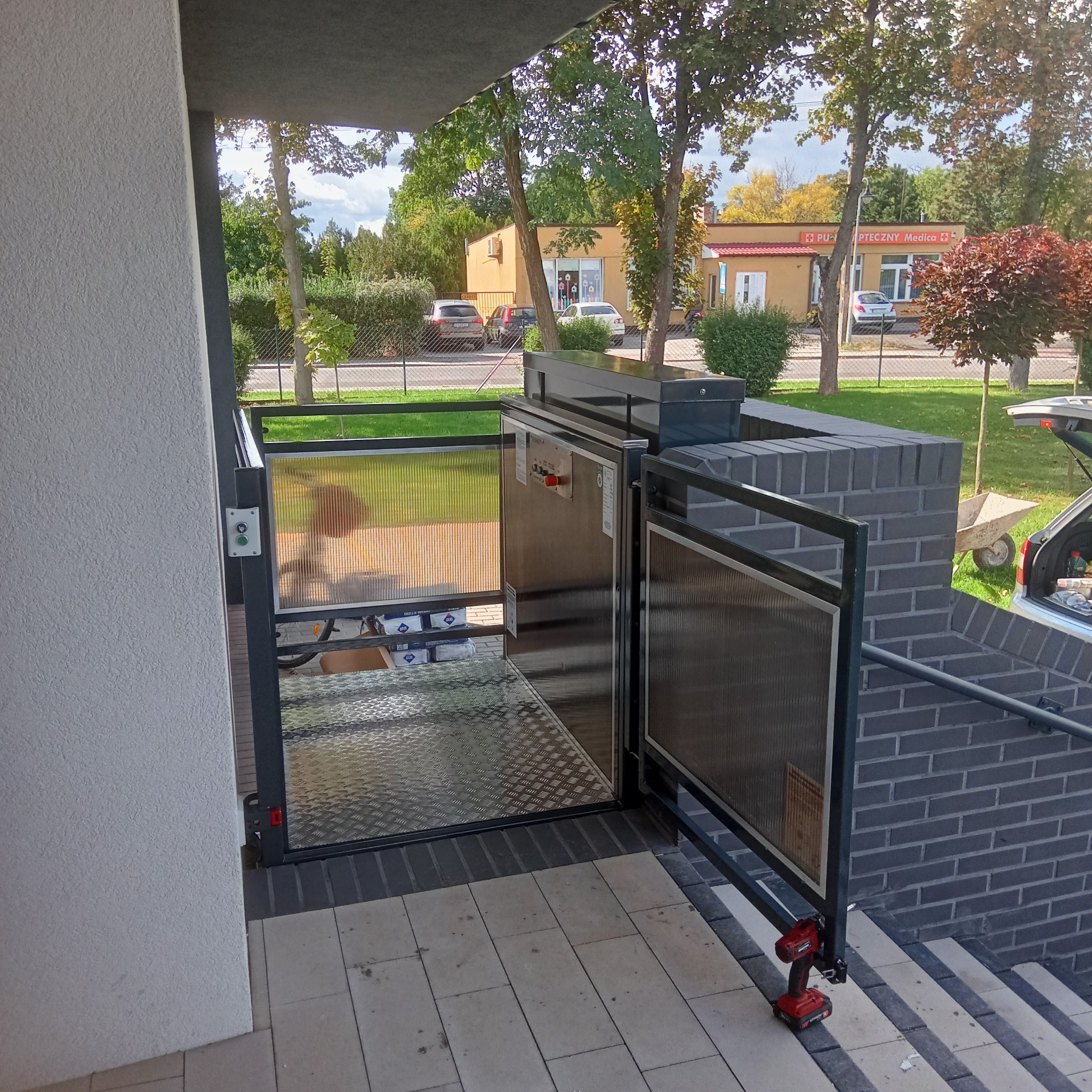 Platforma schodowa dla osób niepełnosprawnych, zamontowana przy wejściu do budynku, z widocznymi elementami konstrukcyjnymi i narzędziem do montażu (wiertarko-wkrętarka).