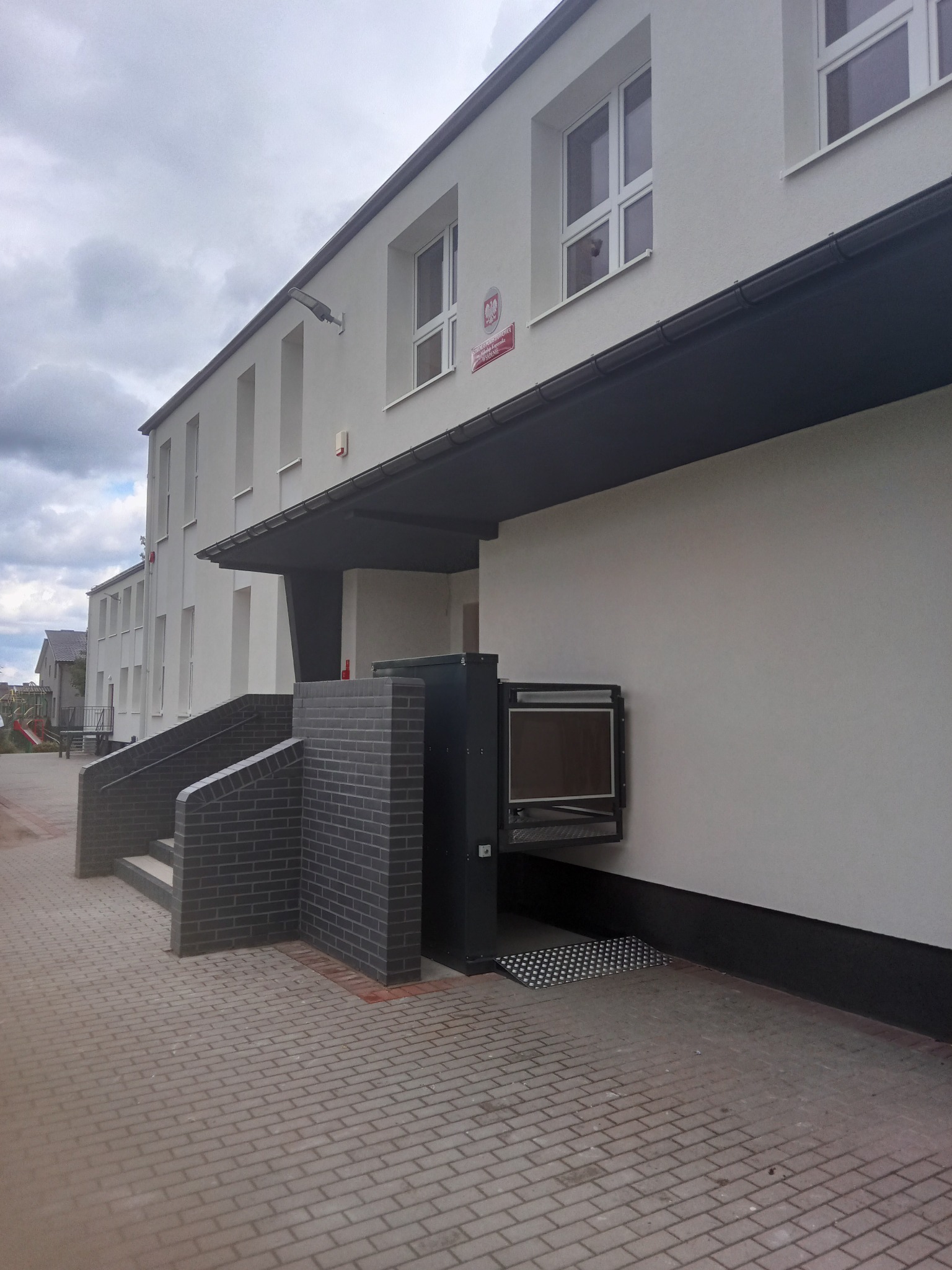 Zewnętrzna platforma podnosząca dla osób niepełnosprawnych, zamontowana przy wejściu do budynku użyteczności publicznej, widoczna z chodnika wyłożonego kostką brukową.